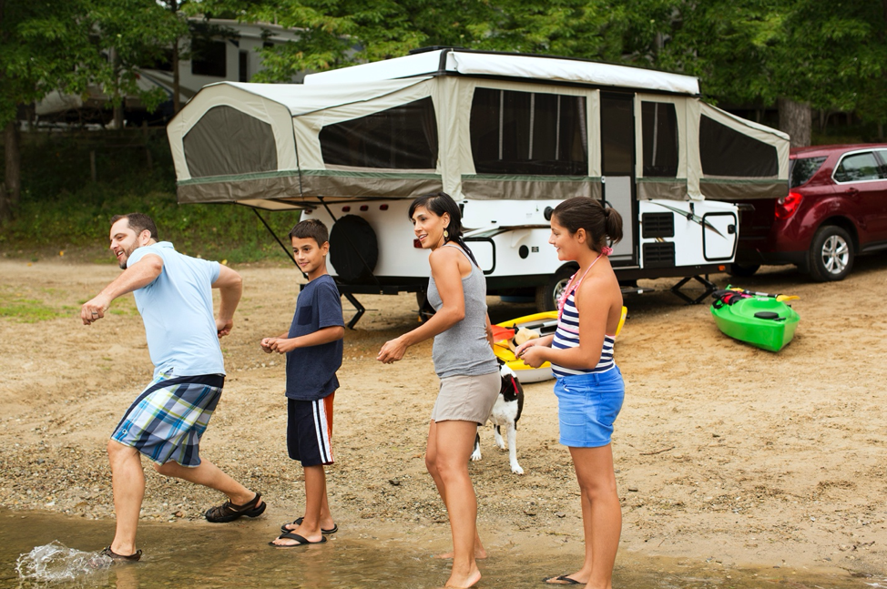 A family playing in a lake with their pop-up camper behind them. 