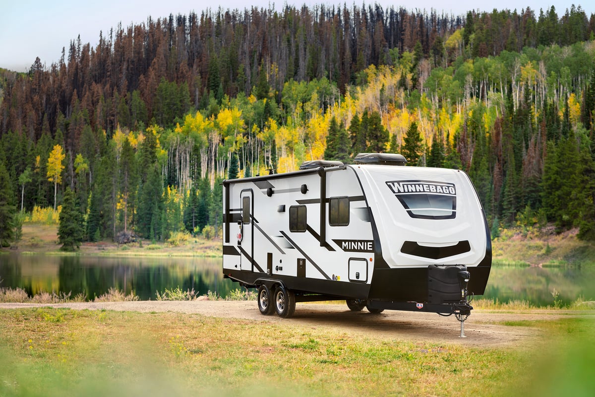 a winnebago minnie travel trailer parked near a lake with mountains and trees in the background