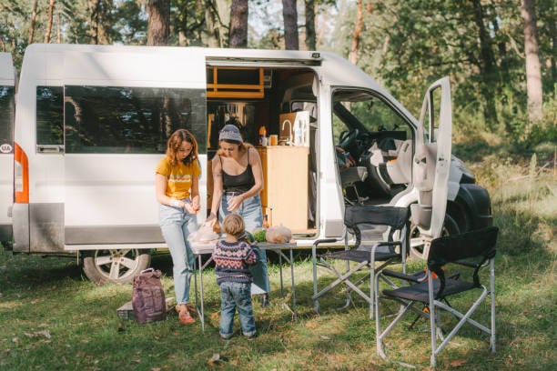 Two women and a child prepare a meal on a small table outside their RV